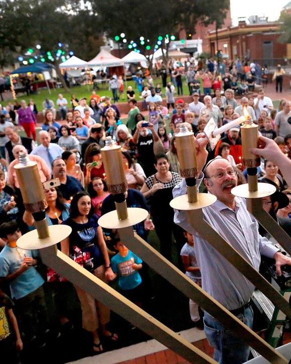 A crowd gathered outdoors at dusk around a large menorah as its lights are lit